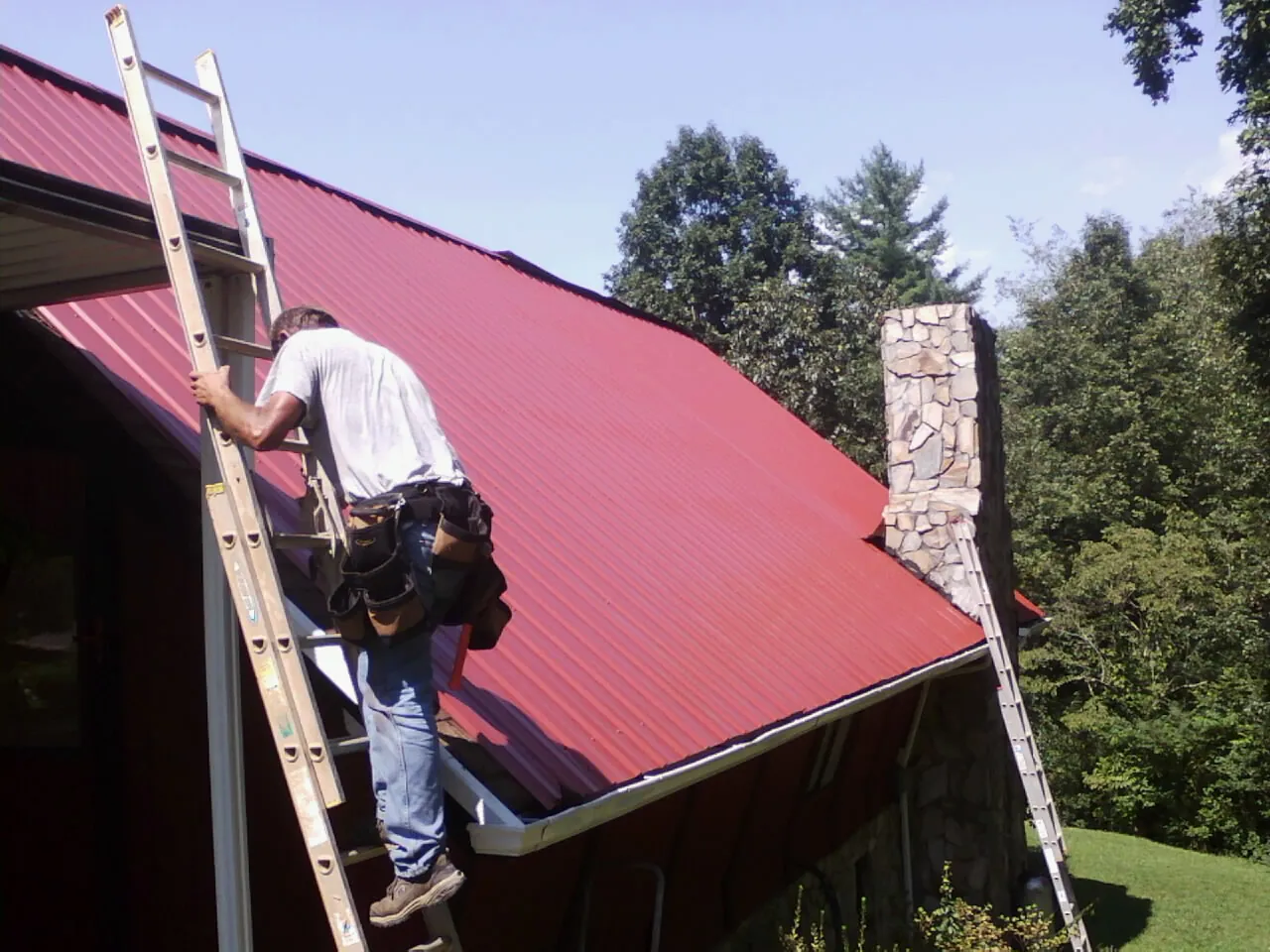 Roofer on a ladder beside a red metal roof and stone chimney — Soffit & Fascia Repair by Roofing Brothers in Lynn
